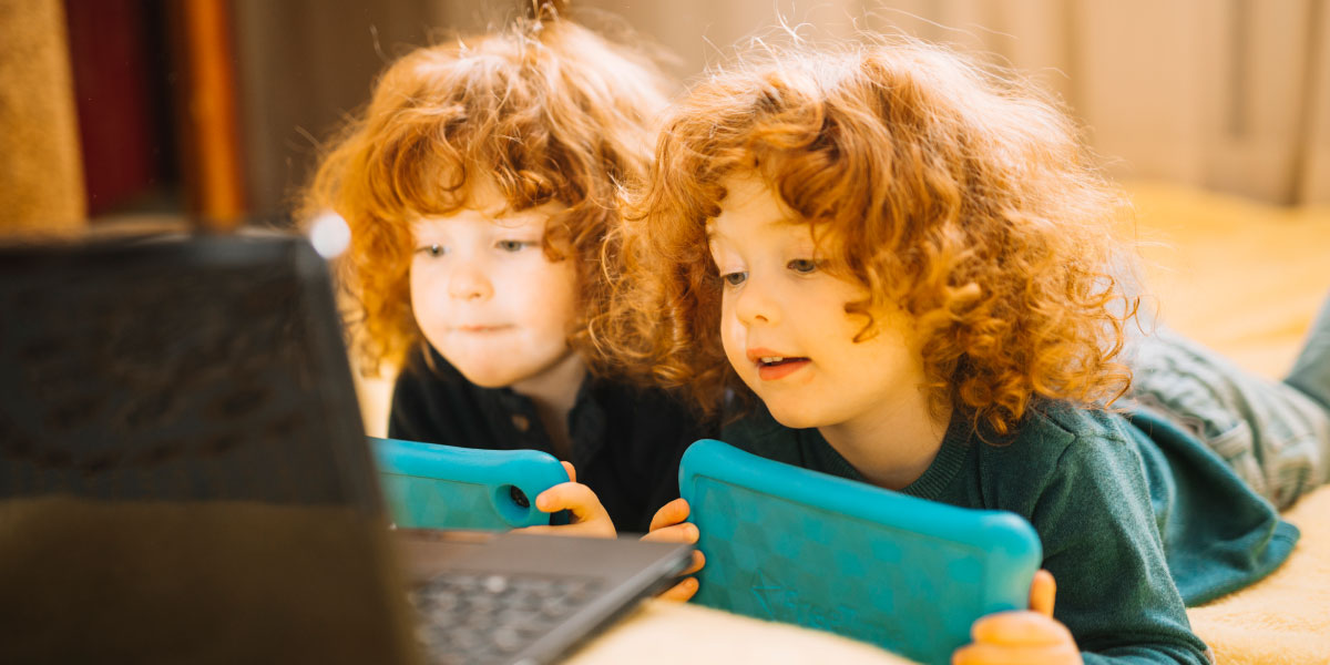 Twins both holding tablets laying on the bed and looking at a laptop